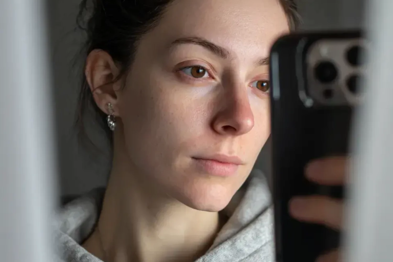 KI-generiertes Bild: Nahaufnahme einer jungen Frau mit dunkelbraunem, locker hochgestecktem Haar und braunen Augen, die ein Spiegel-Selfie mit einem schwarzen Smartphone macht. Das Gerät verdeckt einen Teil ihres Gesichts. Sie trägt einen grauen Hoodie und einen kleinen silbernen Ohrstecker. Der Ausdruck ist ruhig und unaufgeregt. Das Bild ist durch den Spiegelrahmen gerahmt, der links und rechts leicht unscharf in den Vordergrund ragt. Die Lichtstimmung ist natürlich und weich.
